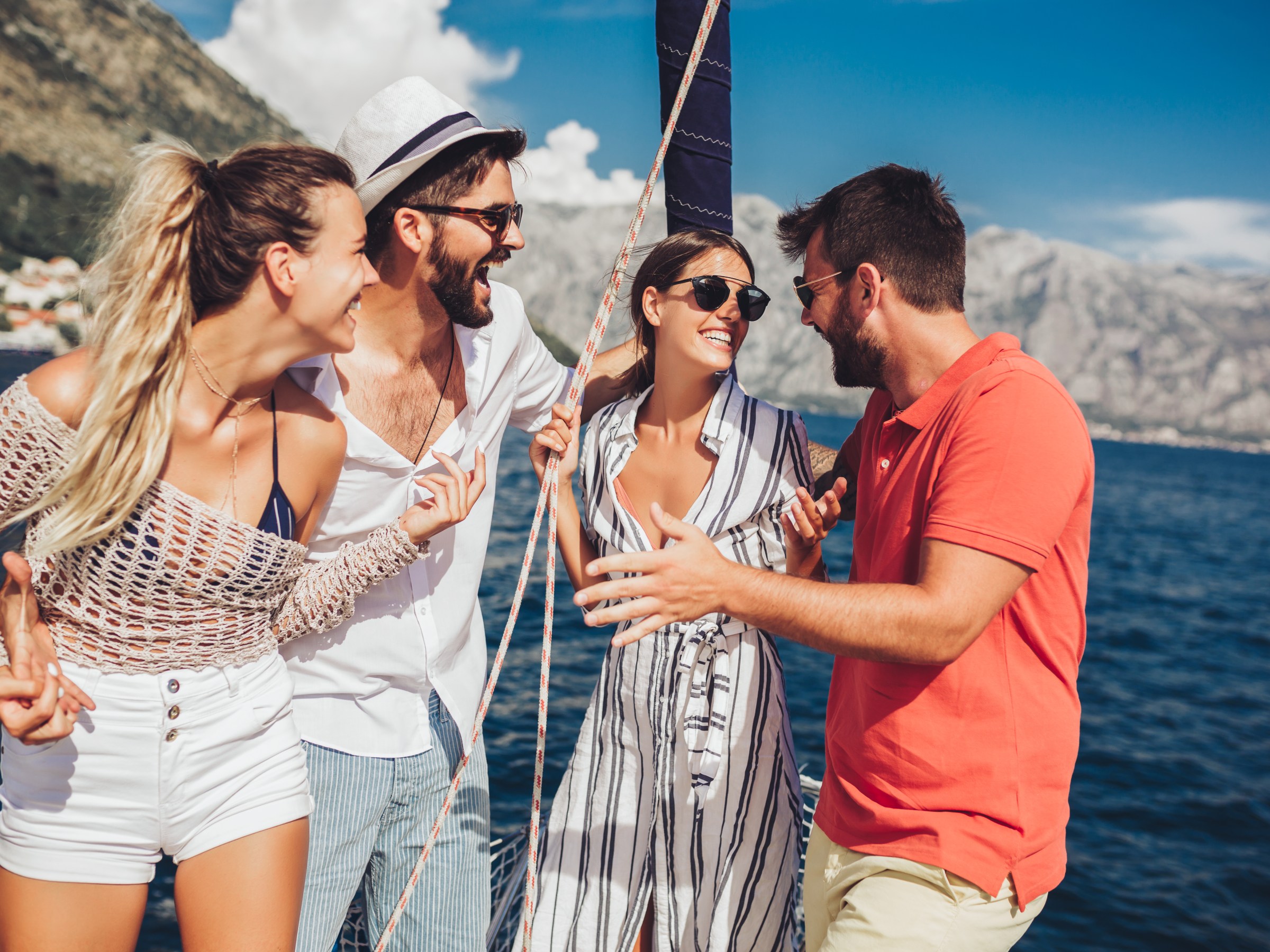 Four friends laughing and chatting on a boat in sunny weather with mountains in the background.