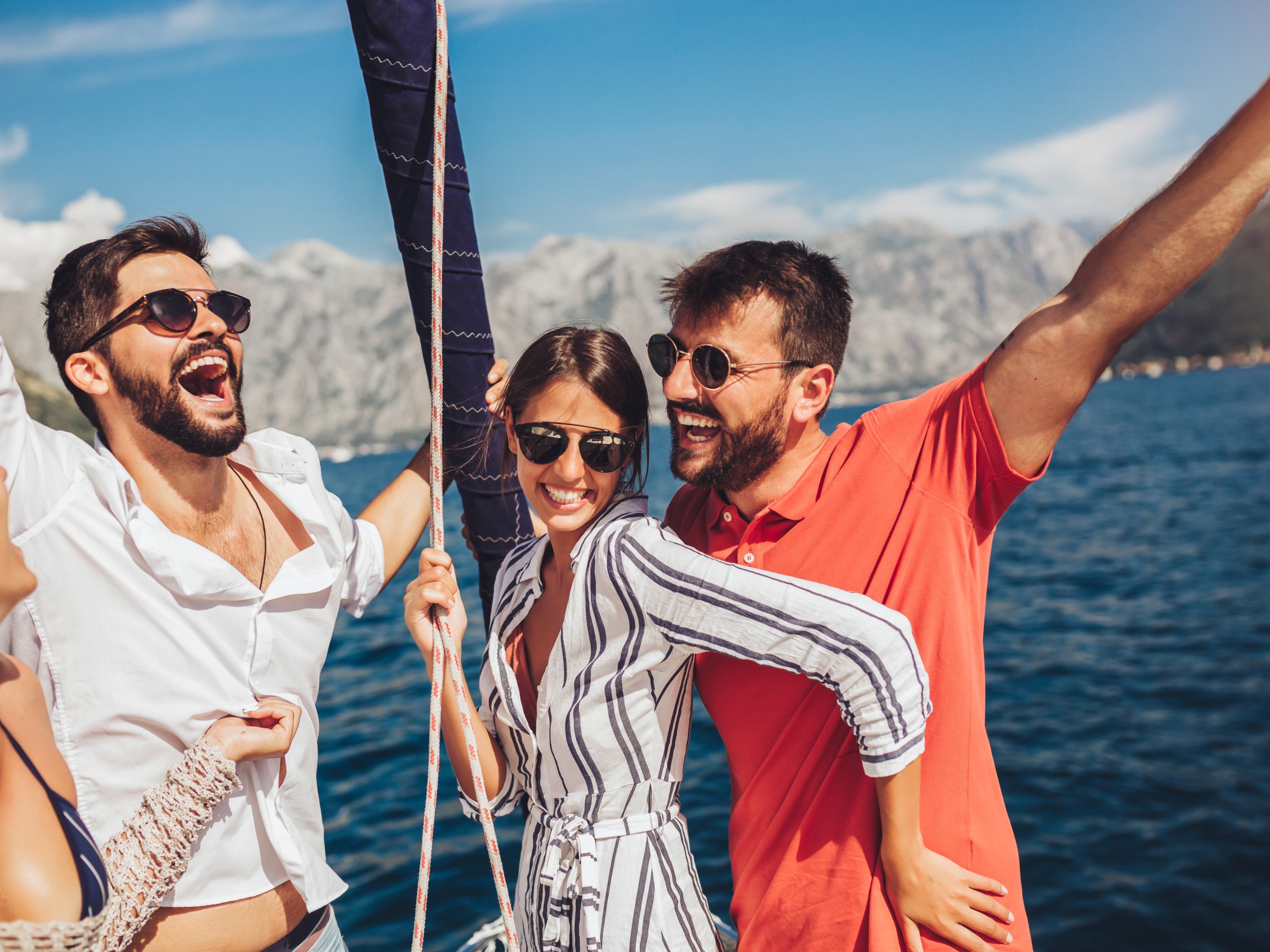 Group of friends laughing on a sailboat, with mountains and water in the background.