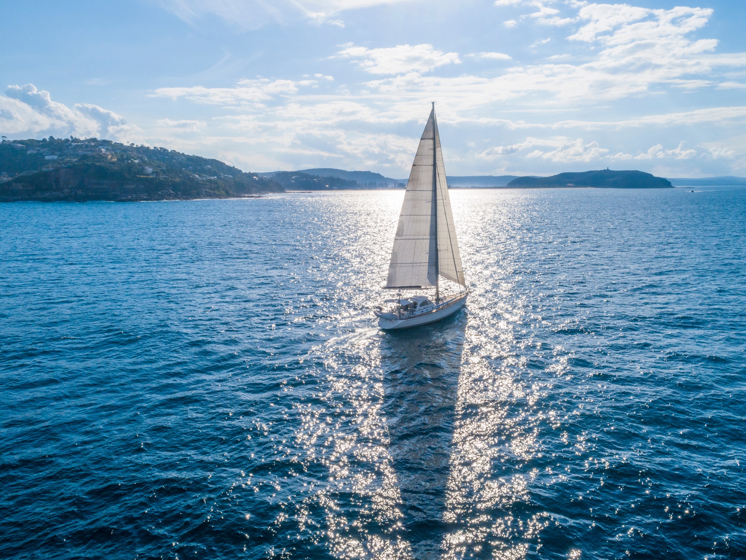 Sailboat on a sparkling blue sea with distant hills under a partly cloudy sky.