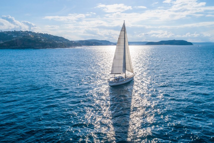 Sailboat on a sparkling blue sea with distant hills under a partly cloudy sky.