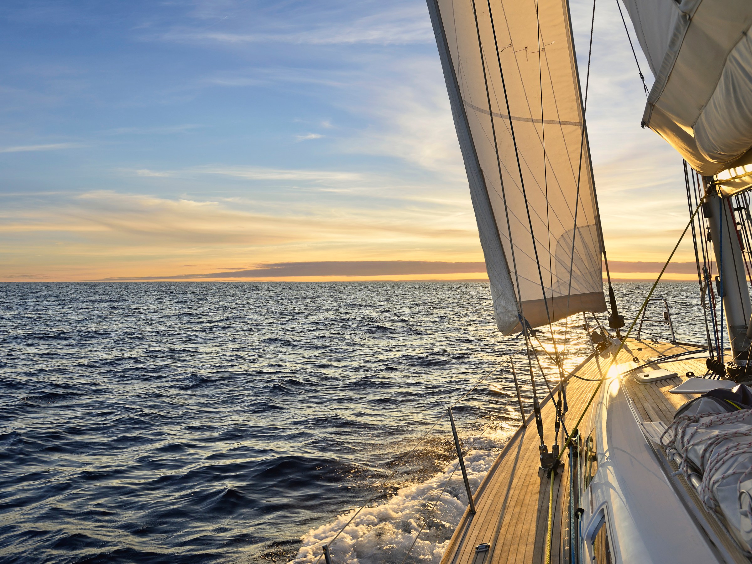 Sailboat cruising on open sea during sunset with sails unfurled and sunlight reflecting on water.