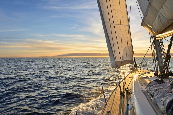Sailboat cruising on open sea during sunset with sails unfurled and sunlight reflecting on water.