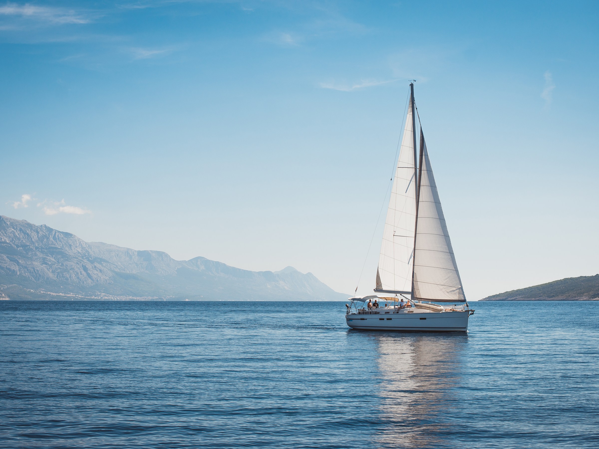 Sailboat on calm sea with mountains and clear blue sky in the background.