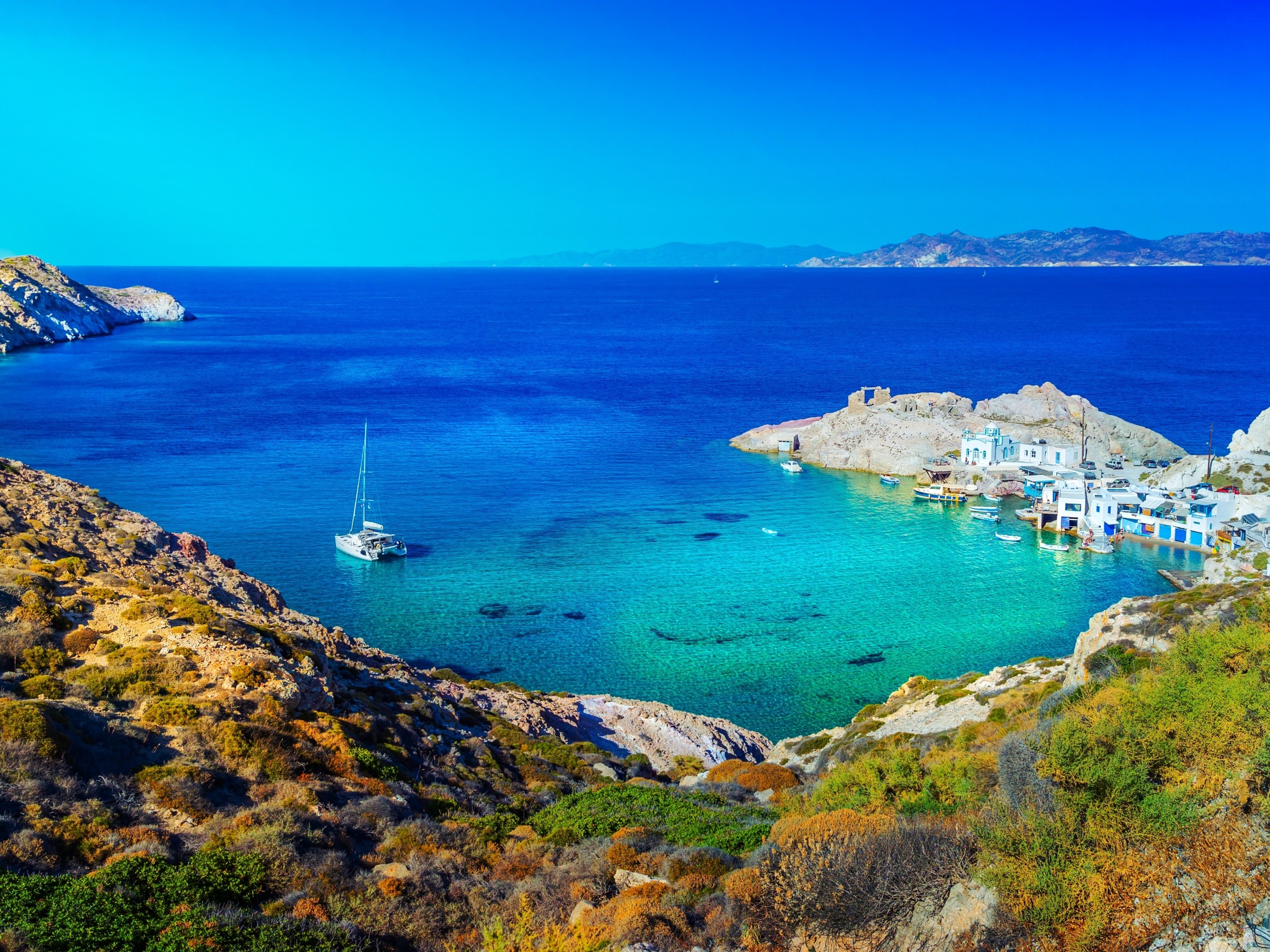 Scenic view of a bay with turquoise water, sailboat, and coastal village under a clear blue sky.