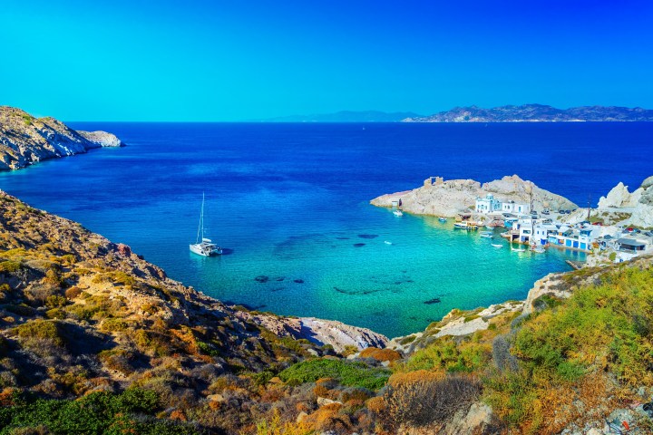 Scenic view of a bay with turquoise water, sailboat, and coastal village under a clear blue sky.