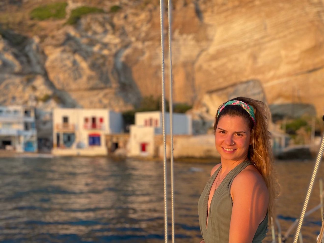 Woman on a boat smiling with rocky hills and buildings in the background at sunset.