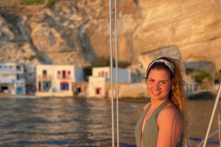 Woman on a boat smiling with rocky hills and buildings in the background at sunset.